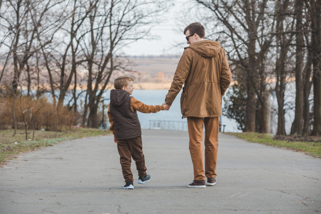 father with son walking by park holding hands. rear view